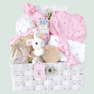 Newborn baby girl gifts in a white hamper basket with organic pink baby clothing and bunny soft toy.