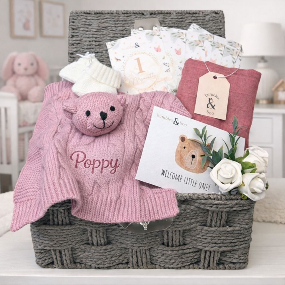 Dusky pink gift hamper for a new baby girl with gifts including teddy comforter, packed in a wicker basket