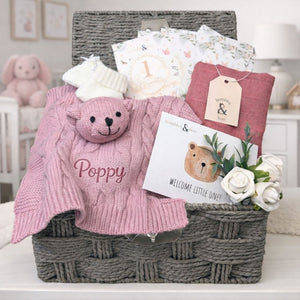 Dusky pink gift hamper for a new baby girl with gifts including teddy comforter, packed in a wicker basket