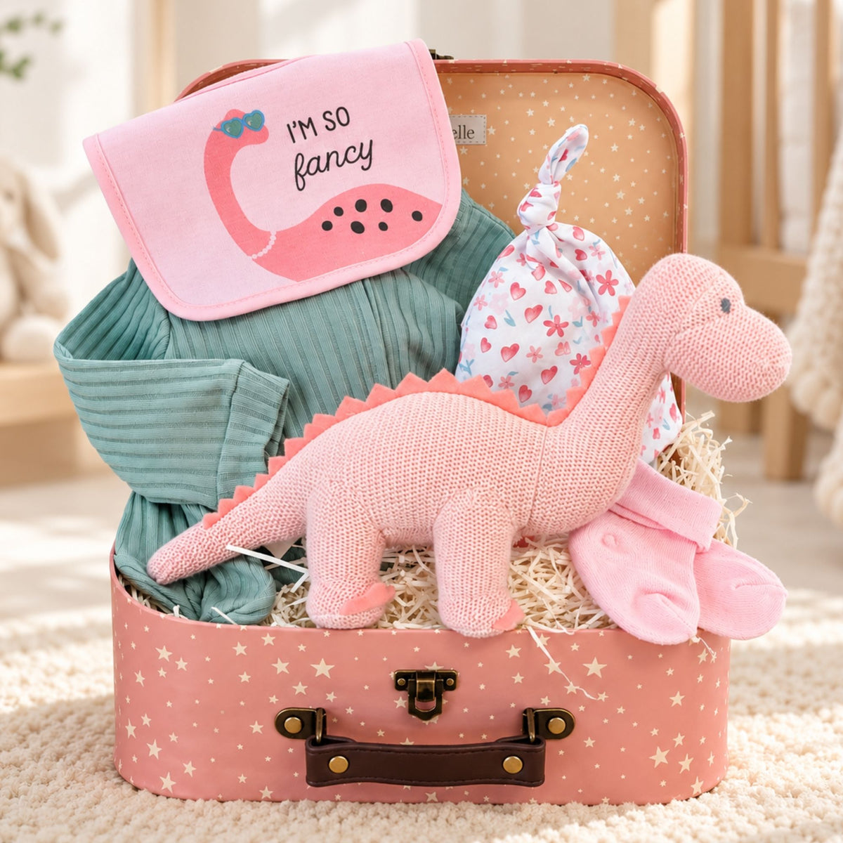 A baby girl gift set arranged inside a pink suitcase with star print. The set includes a pink dinosaur plush toy, a teal ribbed long-sleeve sleepsuit and a pink and white heart-patterned hat and bib set