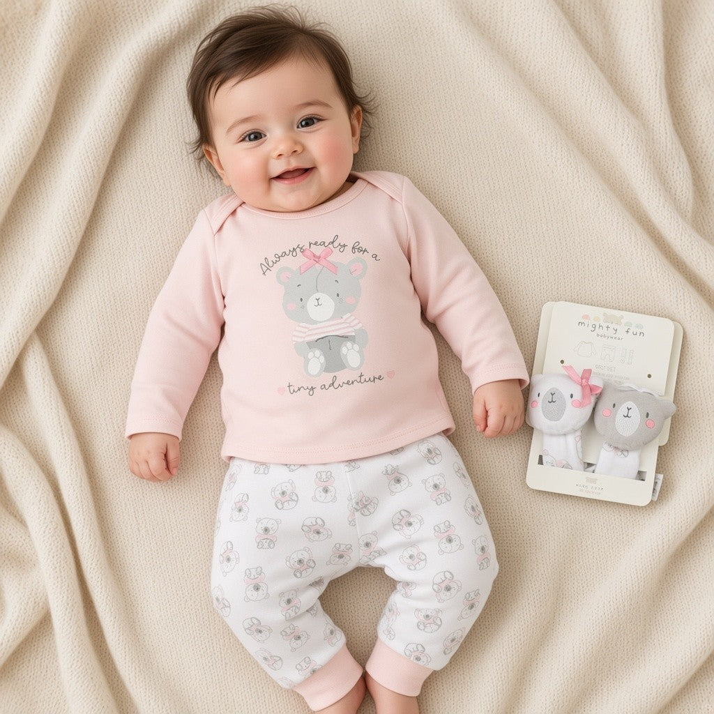 Baby wearing pink bear top and white bear-print trousers, with two wrist rattles beside her on a cream blanket.