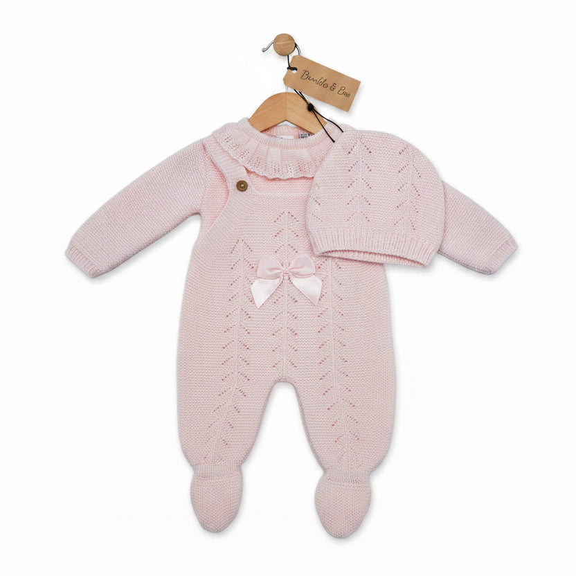A pink baby knit clothing set including a footed one-piece jumpsuit, a matching bonnet, and a small hat, displayed on a white background. The jumpsuit features a textured cable-knit pattern, a ruffled collar, and a small bow at the chest.