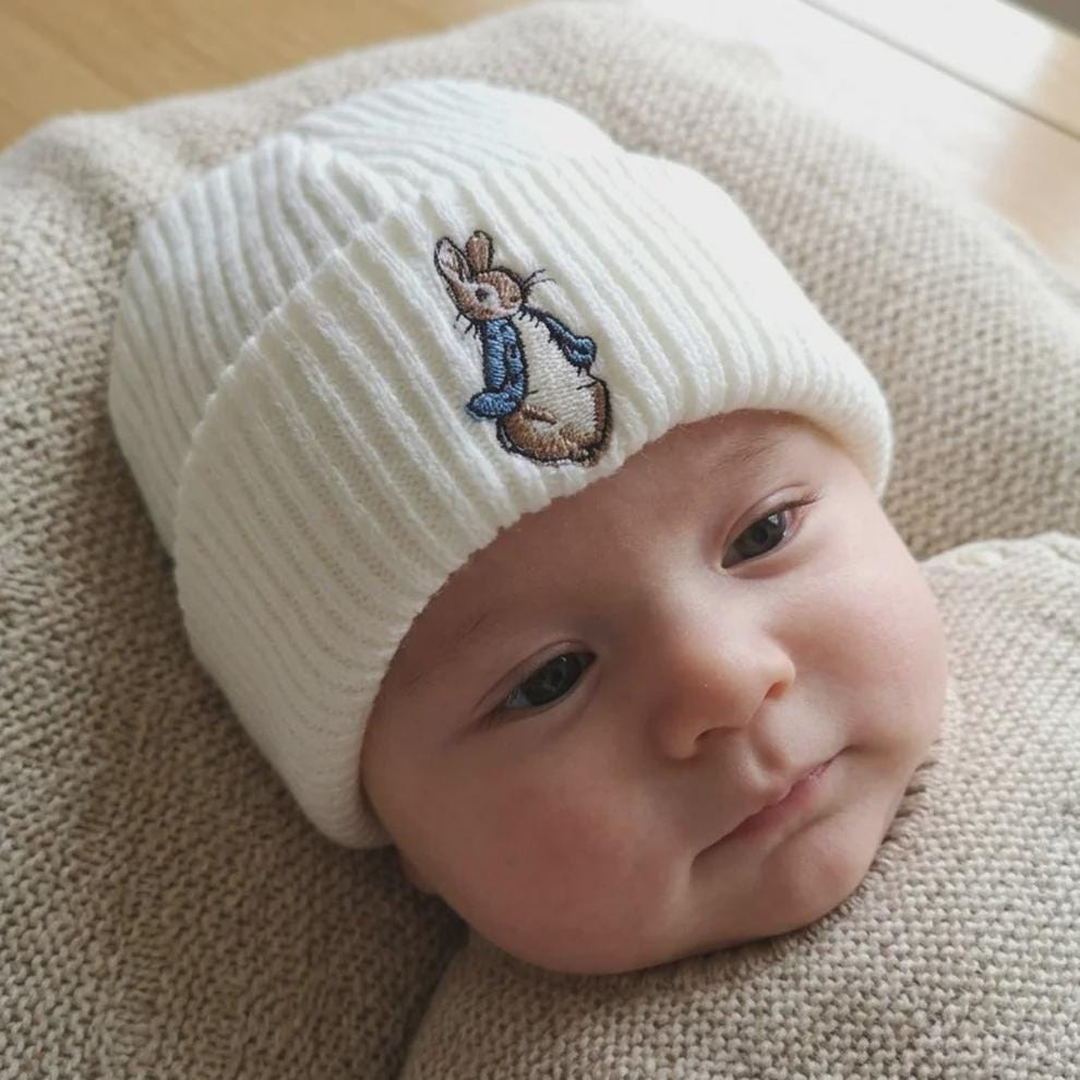 Baby wearing a white ribbed knitted hat with a blue-jacketed bunny embroidered on the cuff.
