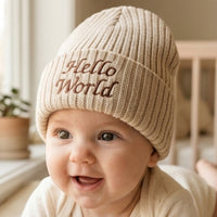 A close-up of a smiling baby wearing a beige ribbed knit beanie with 'Hello World' embroidered on the front in brown lettering, set in a softly lit indoor environment.