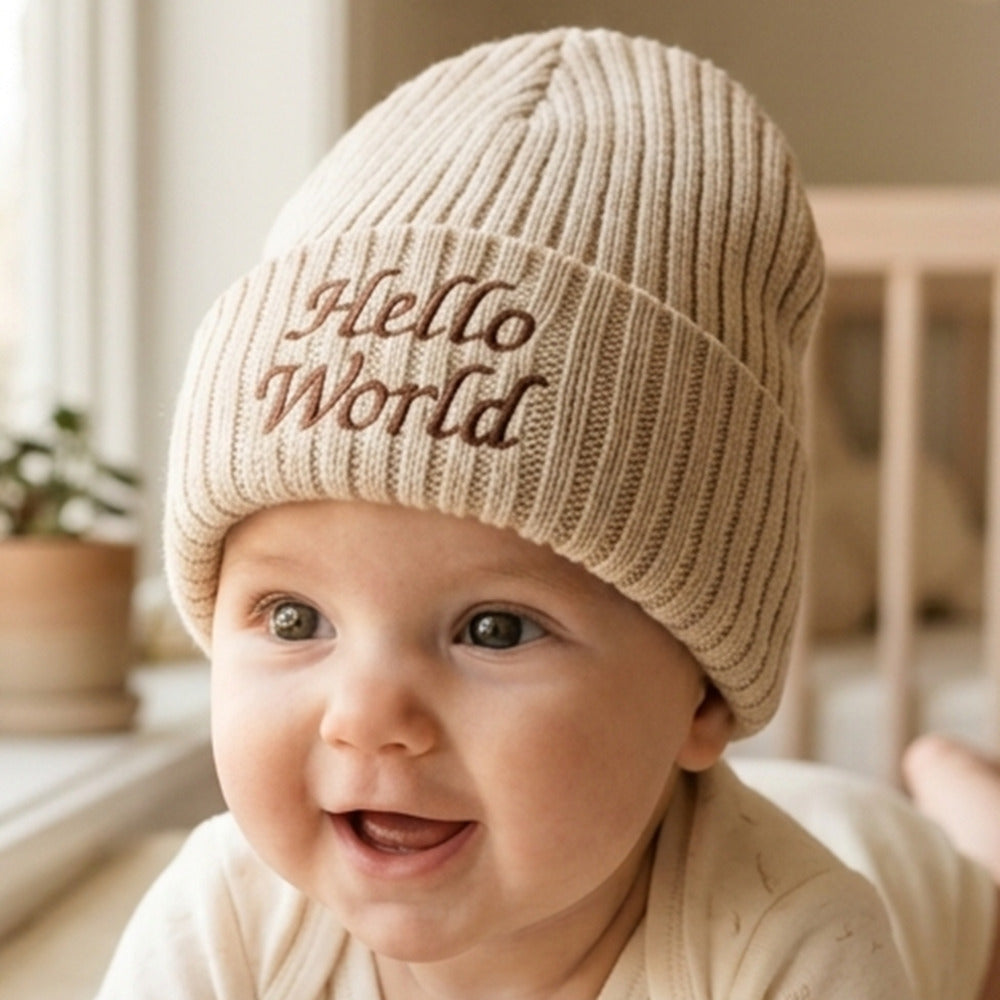 A close-up of a smiling baby wearing a beige ribbed knit beanie with 'Hello World' embroidered on the front in brown lettering, set in a softly lit indoor environment.