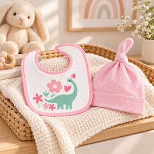 A baby gift set featuring a white bib with pink trim and a green dinosaur design, paired with a soft pink knotted beanie hat