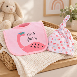 A pink baby bib with a pink dinosaur and the text 'I'M SO fancy' is displayed next to a matching pink and white floral baby hat 
