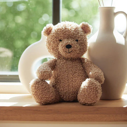 Teddy bear sitting on a windowsill with a vase and decorative grasses.