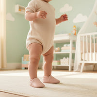 Baby standing wearing blush pink baby shoes in soft vegan leather, lightweight baby booties styled with neutral newborn baby clothes.