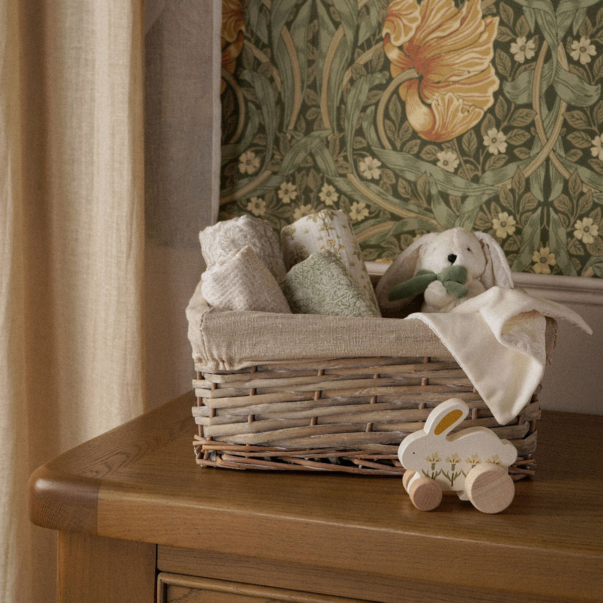 Cream bunny comfort blanket hanging over a basket edge in a floral nursery setting