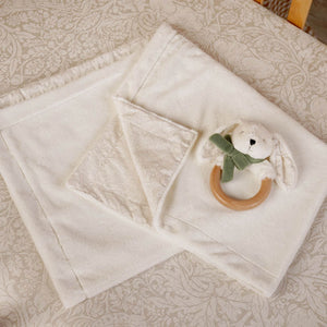 Cream baby blanket with a folded patterned corner and bunny ring teether on a patterned surface.
