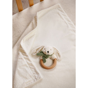 Plush cream baby blanket draped over a chair with a bunny wooden ring teether resting on it.