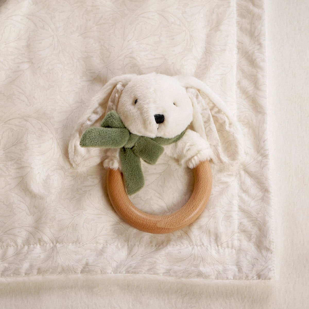 Close view of a plush bunny teether with sage bow and wooden ring on the blanket’s patterned reverse.