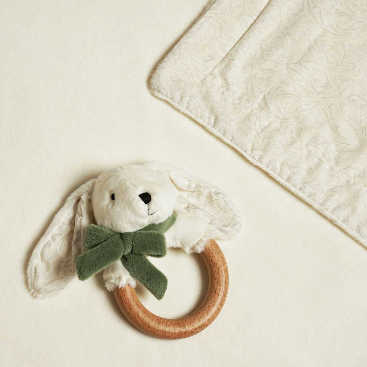 Bunny ring teether beside a turned-back corner showing the cream blanket’s leafy patterned reverse.