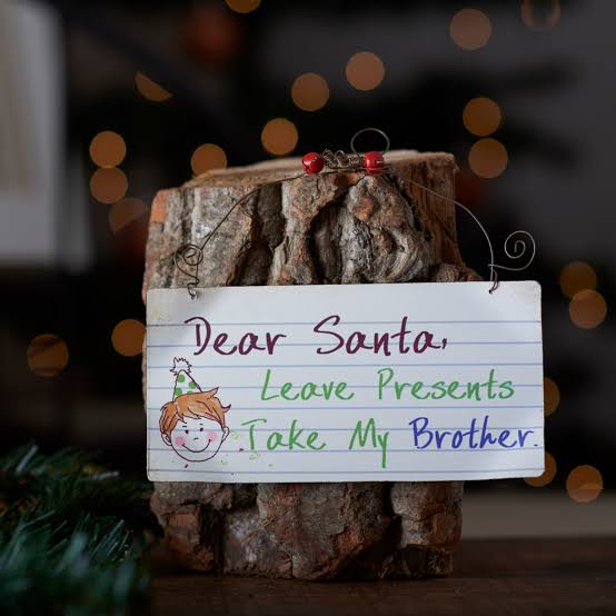 Dear Santa Leave Presents Take My Brother sign with coloured lettering, wire hanger and red bells on a log.