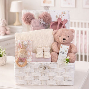 Gifts for a new baby girl in a white hamper basket with teddy bear, knitted hat and cosy baby blanket