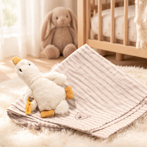 Cream Gilda goose rattle on a striped cotton burp cloth, displayed beside a wooden cot in a nursery.