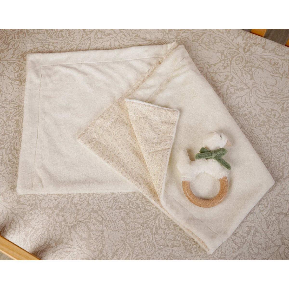 Cream baby blanket with a folded patterned corner and goose ring teether on a patterned surface.
