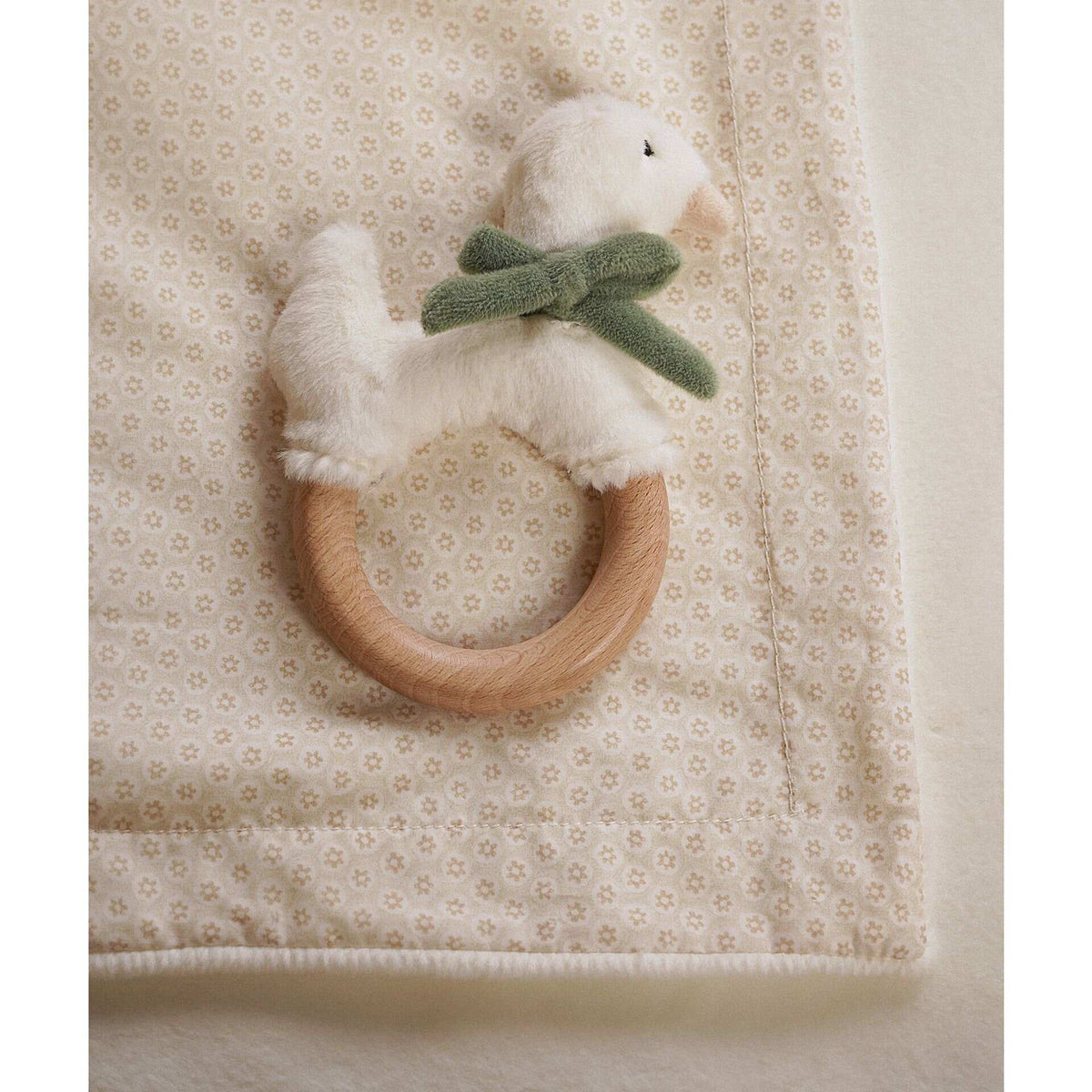 Close view of a plush goose with sage bow and wooden teething ring on the blanket’s floral reverse.