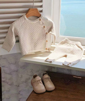 Ivory honeycomb knit baby outfit with asymmetric wooden buttons displayed on nursery windowsill with matching trousers.