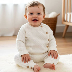 Ivory knitted baby clothes set with honeycomb knit top and matching trousers worn by baby sitting on soft nursery rug.