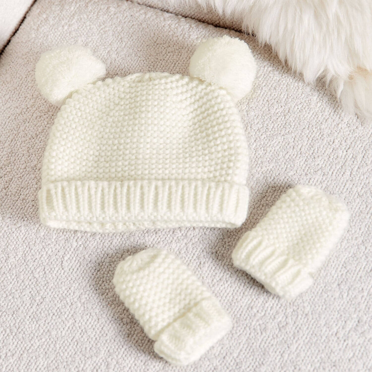 White knitted Katie Loxton hat with two pom-pom ears and matching mittens on a fabric background.