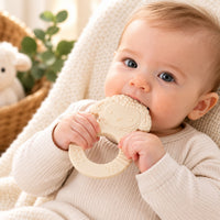 Baby holding and chewing a cream lamb-shaped teether with an open ring grip.