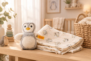 Grey Lio penguin rattle sitting on the folded cream burp cloth with its penguin print.