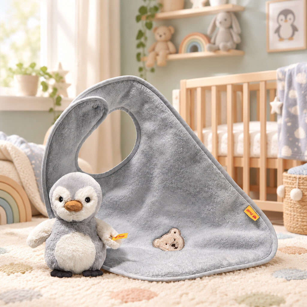 Grey Lio penguin rattle beside a matching cotton bib with teddy bear embroidery, displayed in a nursery.