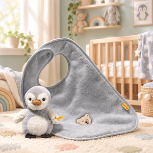 Grey Lio penguin rattle beside a matching cotton bib with teddy bear embroidery, displayed in a nursery.