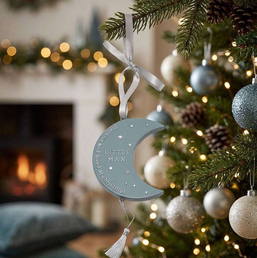 Blue crescent moon Little Man plaque with silver star details and a hanging ribbon beside a Christmas tree.