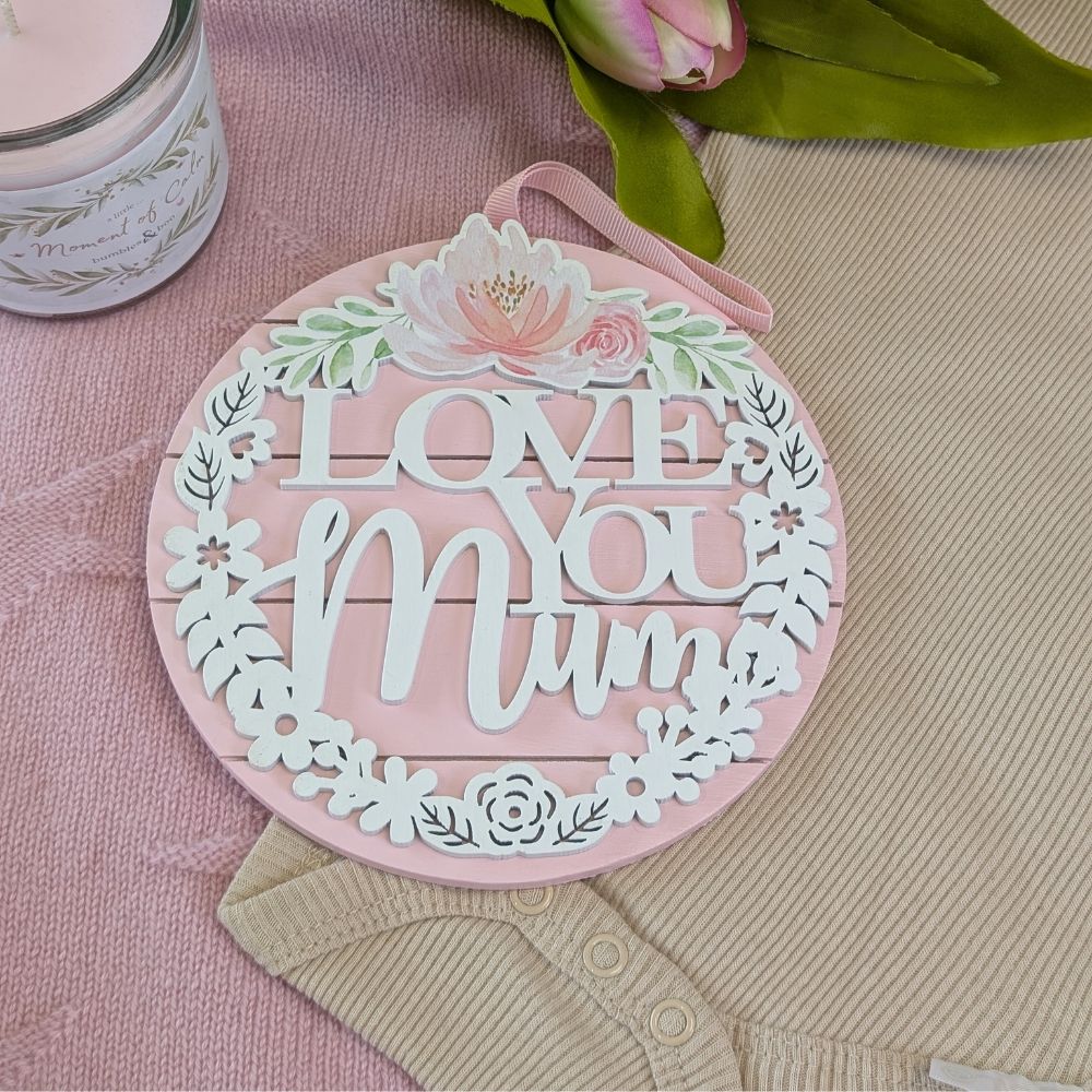 Round pink Love You Mum hanging plaque with white lettering, floral details and a ribbon loop