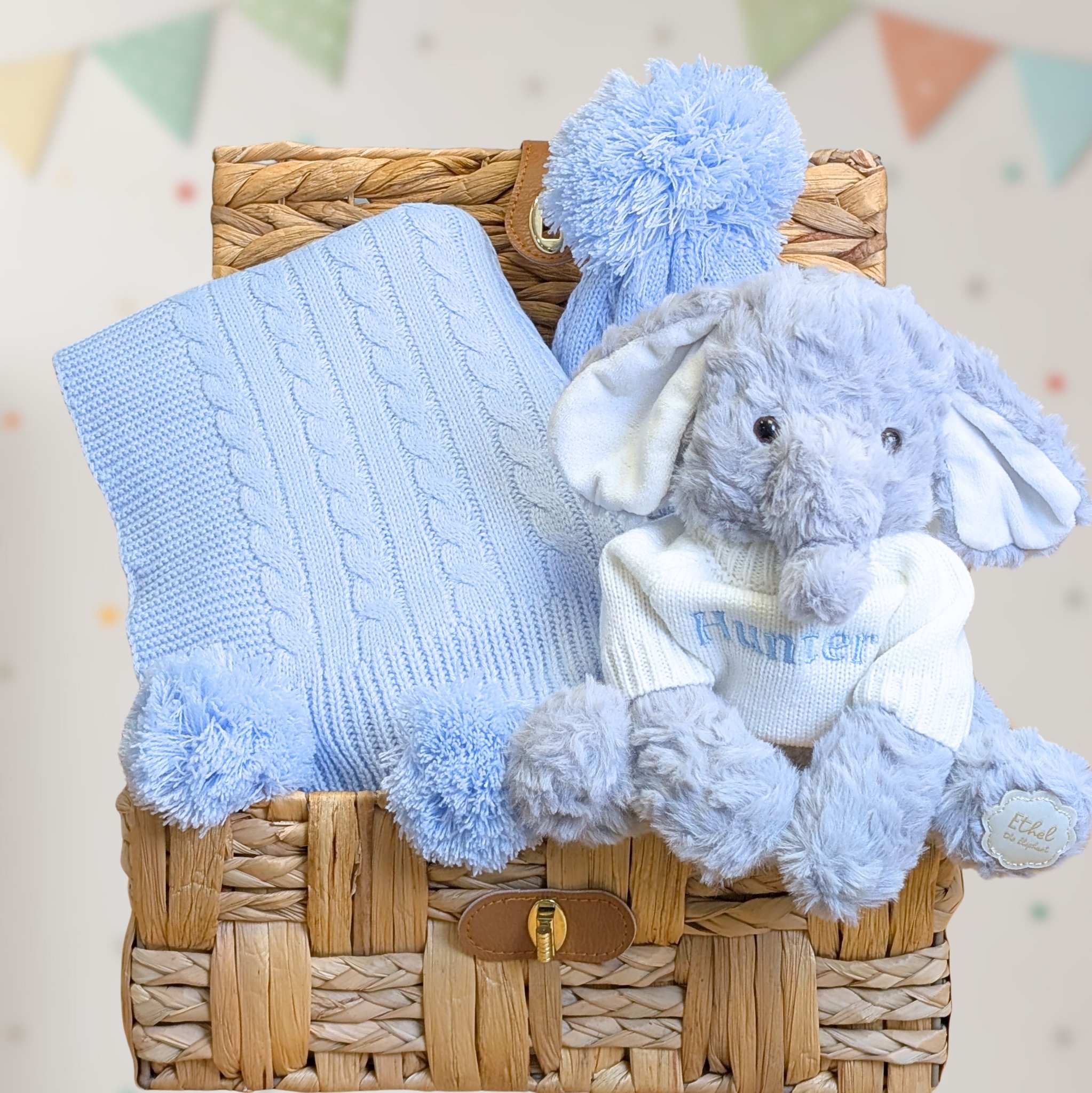 Baby boy gifts set with blue knitted blanket and matching hat plus a plush elephant in a hamper basket.