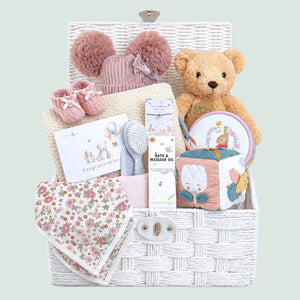 Gifts for a baby girl in a white hamper basket with blanket, teddy bear and an array of presents