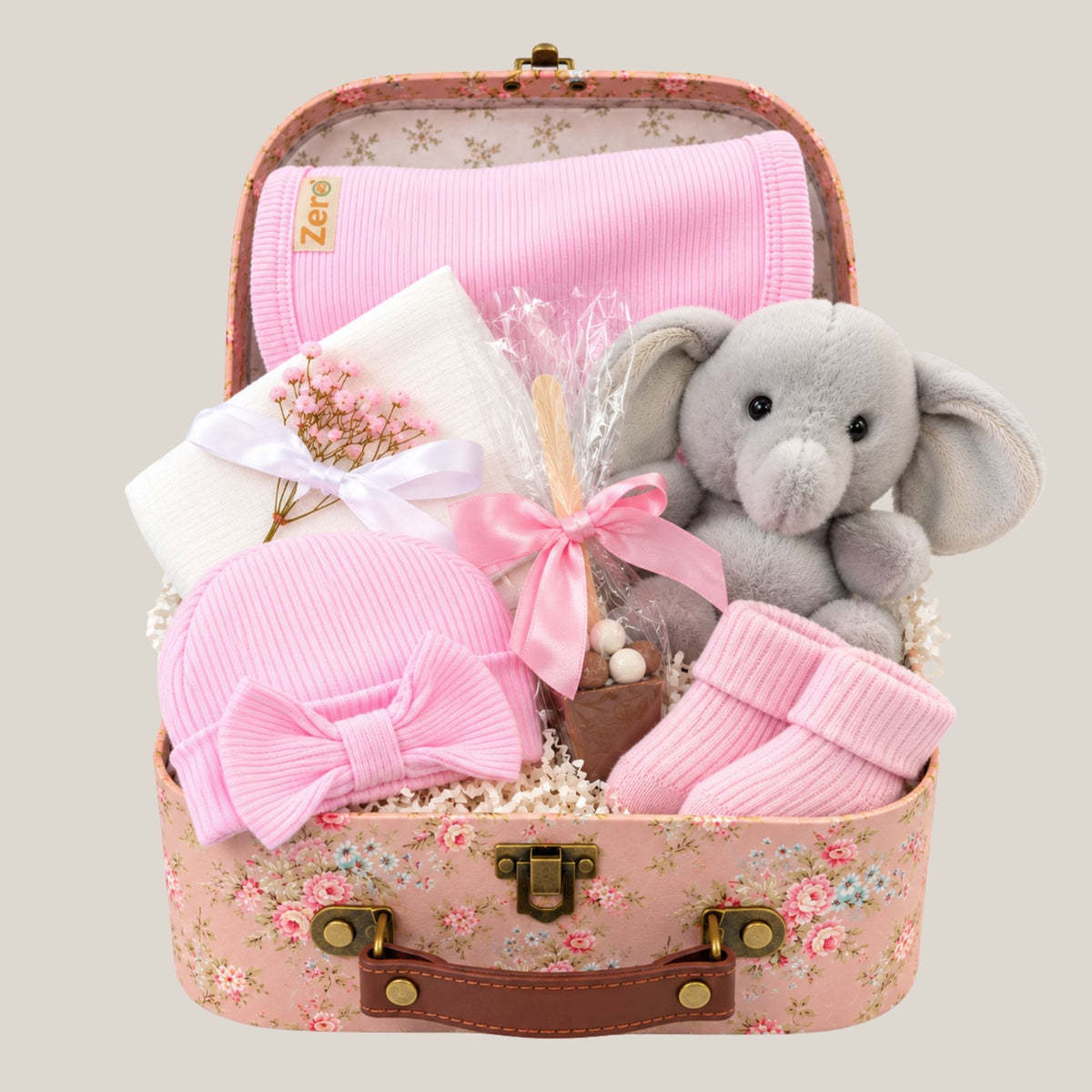 Baby girl gift hamper in a pink floral keepsake trunk with grey elephant soft toy, pink bib, baby hat, socks, white muslin and chocolate stirrer.