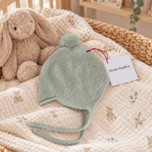Mint green cashmere bonnet with ties and a pom-pom displayed beside a soft rabbit toy.