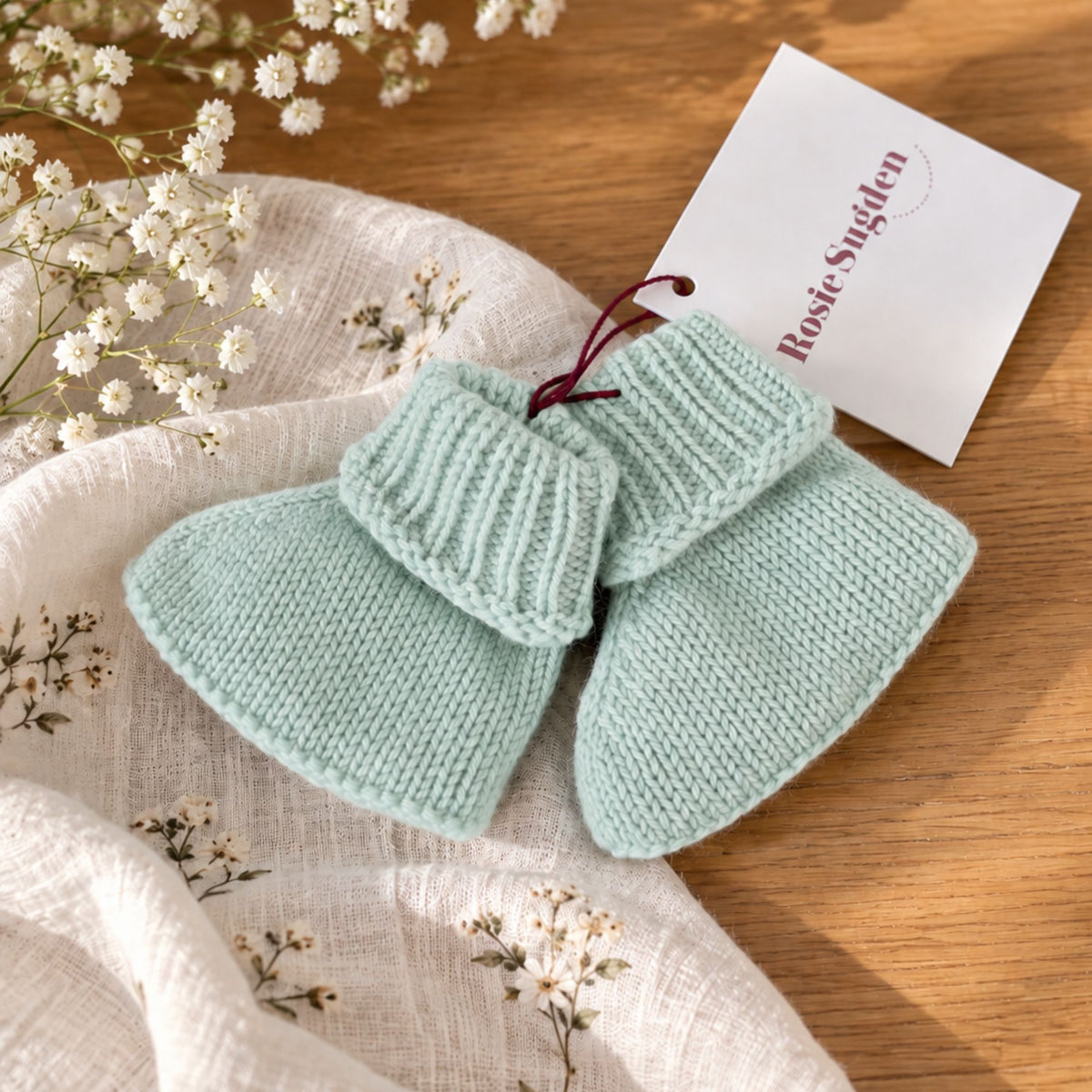 Pair of mint green cashmere baby booties with ribbed cuffs beside a cream blanket and white flowers.