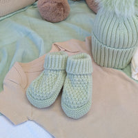 Pair of mint knitted baby booties with folded ribbed cuffs, styled with baby clothes and a hat.