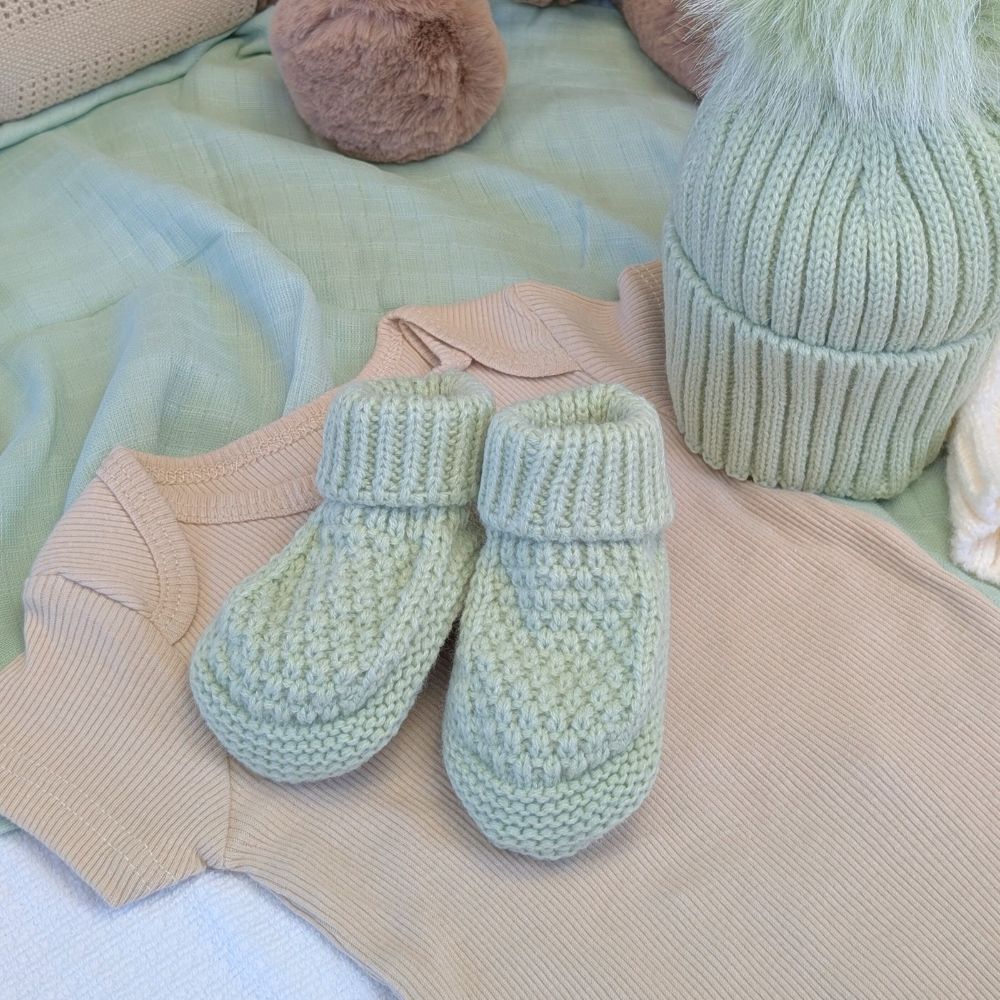Pair of mint knitted baby booties with folded ribbed cuffs, styled with baby clothes and a hat.