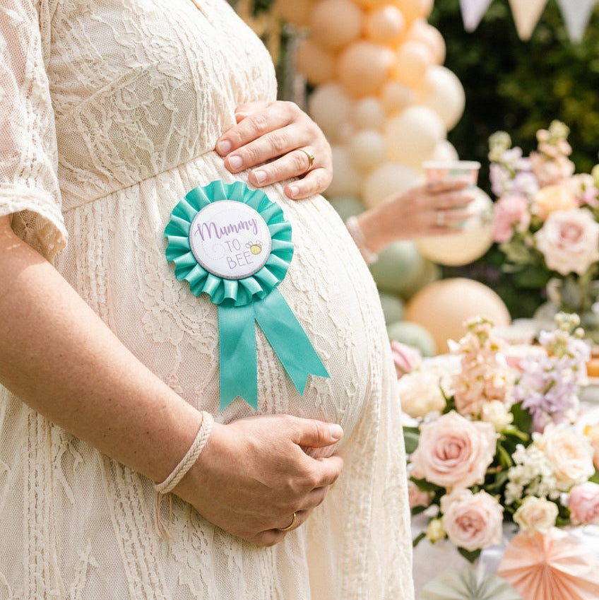 Green Mummy to Bee rosette badge with ribbon tails worn on a cream dress at a baby shower.