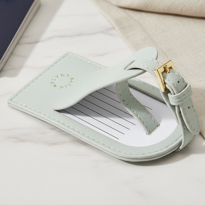 Blue My First Luggage Tag in vegan leather with gold lettering displayed beside suitcase and travel accessories.