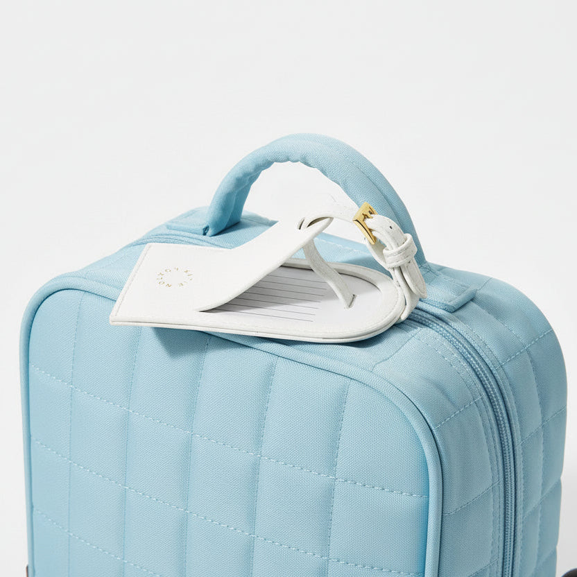 White vegan leather My First Luggage Tag with gold buckle and stitched edge shown on blue travel case.