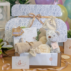 New baby gifts set with teddy bear, blanket, and cards in a hamper box with neutral grey and beige theme.