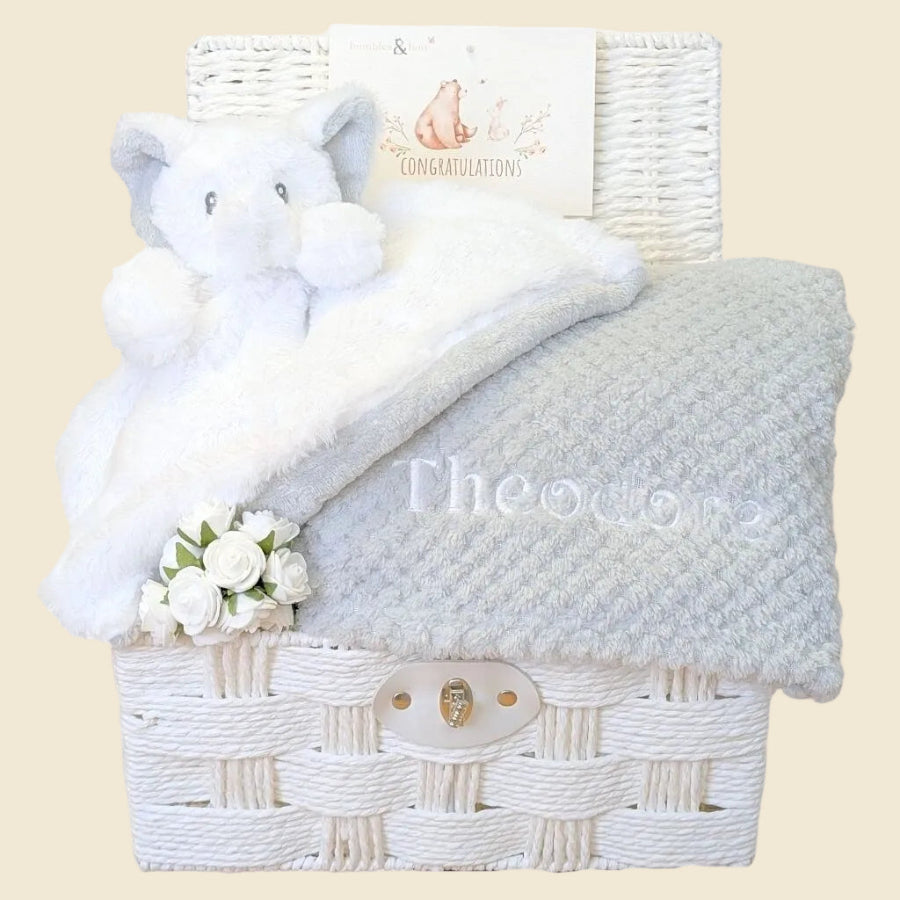 White wicker baby gift hamper basket with gifts including plush toys and a grey blanket 