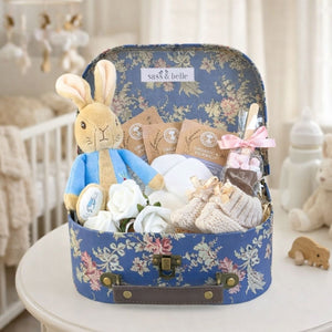 New baby gifts hamper in a blue floral-patterned suitcase. Presents include a Peter Rabbit soft toy and treats for mum.