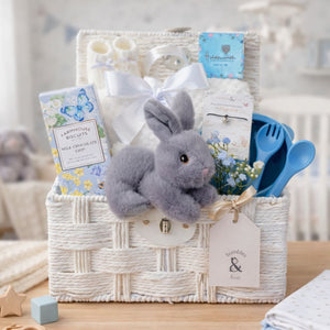 Newborn baby gifts in a white wicker hamper basket with bunny, blanket and feeding bowl.