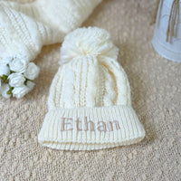 Personalised baby hat in a cream pearl and cable knit design with a large pom pom