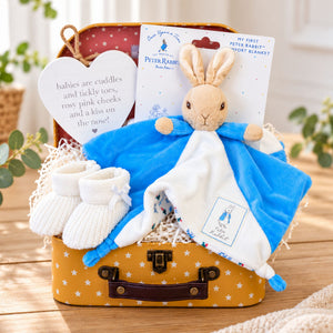 A Peter Rabbit baby gift set arranged in a small yellow suitcase and includes a blue and white blanket with a plush rabbit, white knit booties, a white heart-shaped plaque