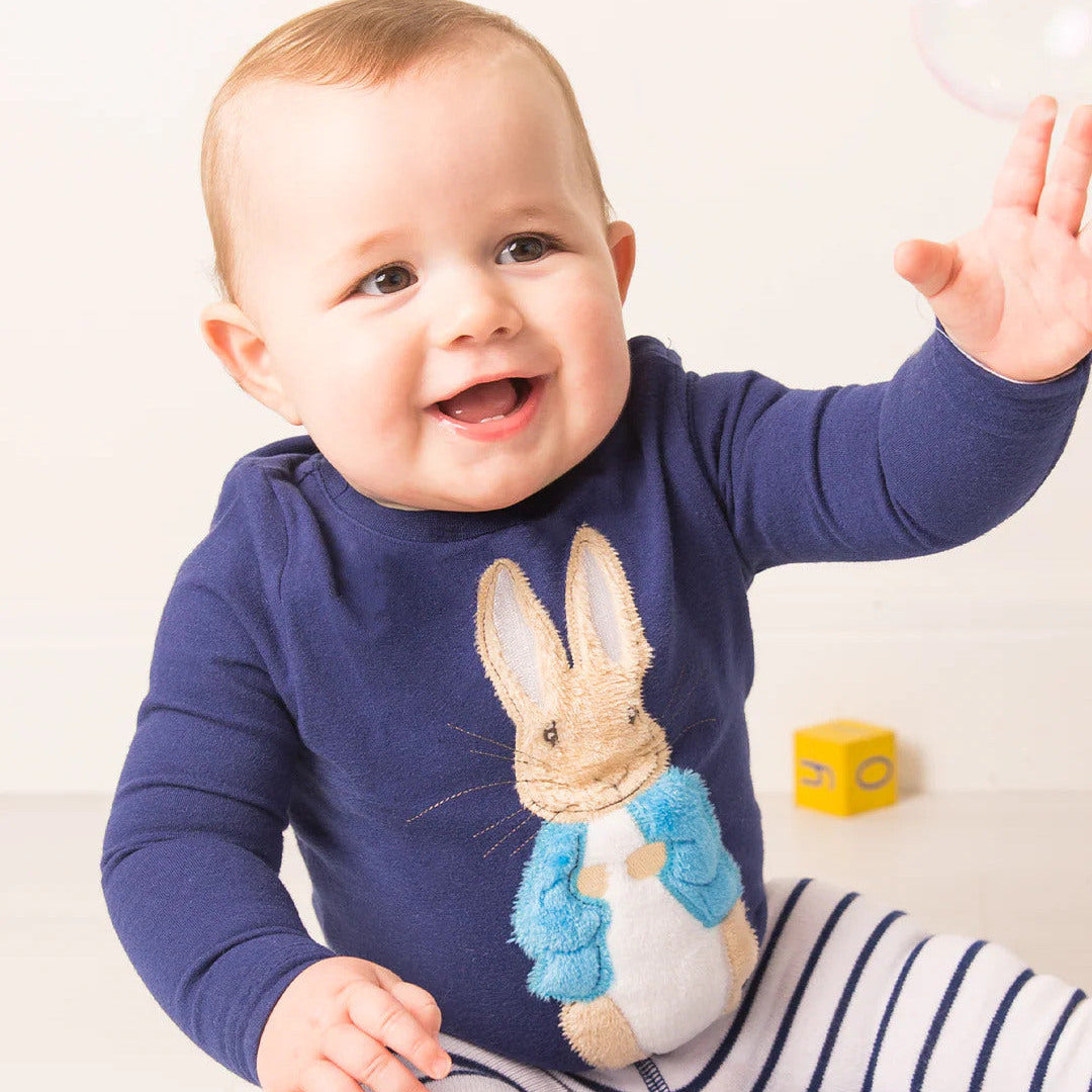 Baby wearing a navy Peter Rabbit long sleeve top while seated on a white surface