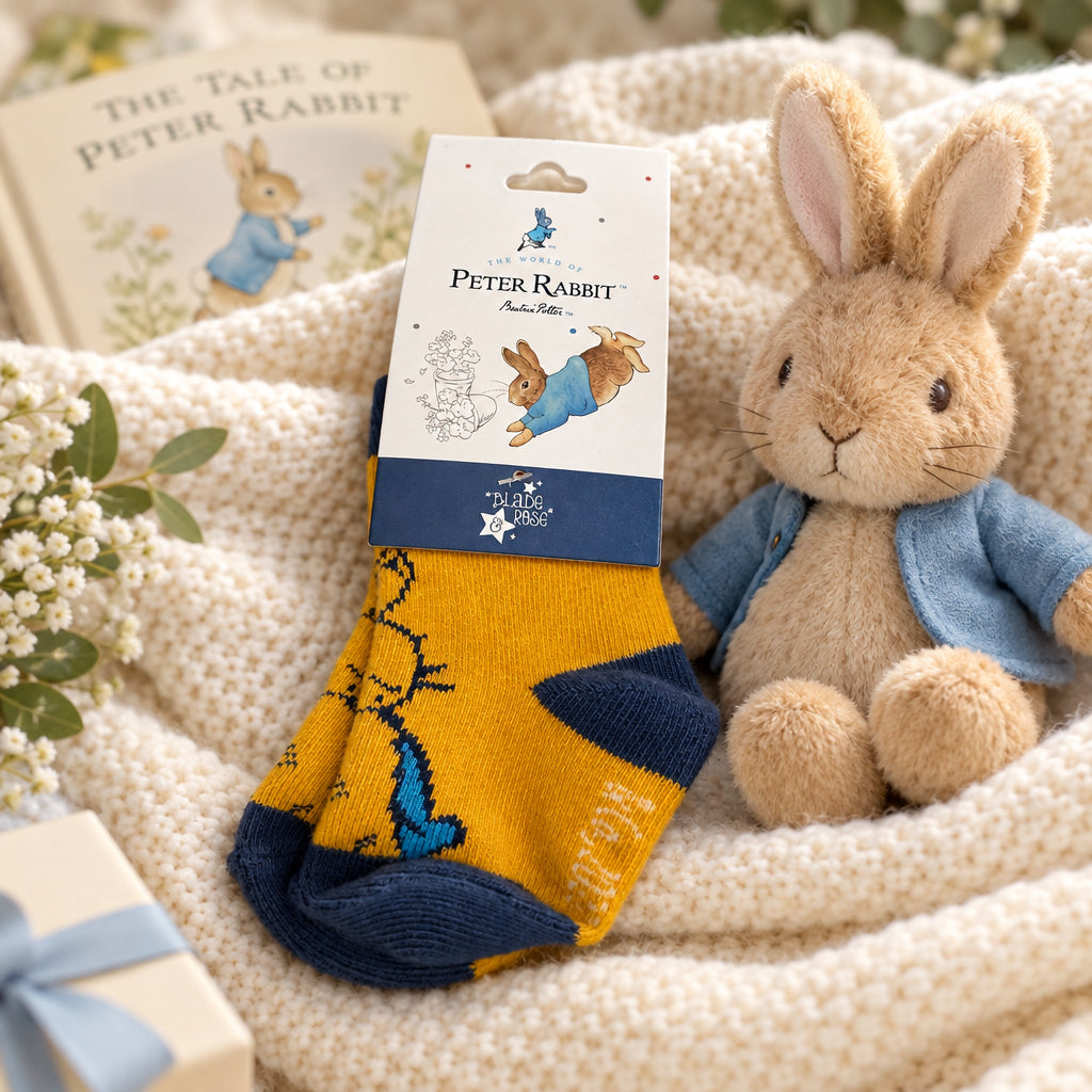 Yellow and navy Peter Rabbit baby socks on a cream blanket beside a rabbit soft toy, with a book in the background.